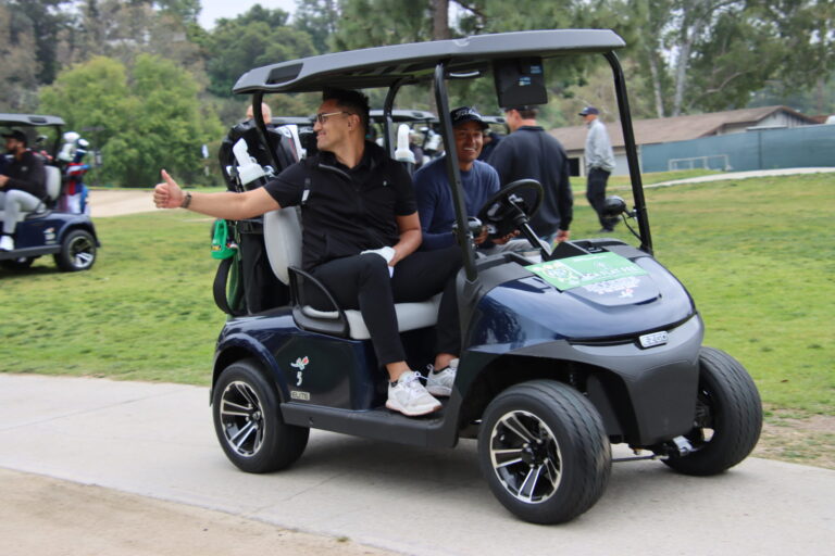 a man in black seated in the passenger seat of a golf cart giving a thumbs up