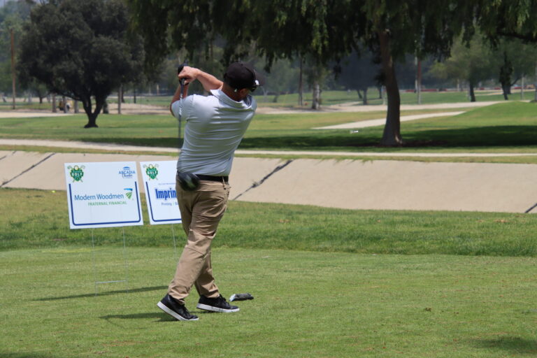 a man in a white shirt and khakis teeing off in front of a few tee signs