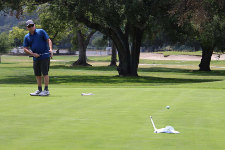 a man in a blue shirt and shorts assessing a putt as the ball rolls towards a hole on a golf green