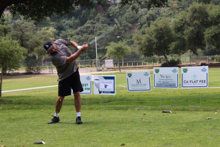 a man in a brown shirt and shorts teeing off in front of a row of tee signs