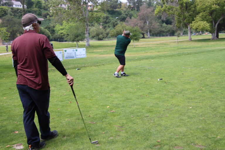 a man in a maroon shirt leaning on a golf club watching a man in green tee off