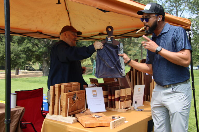 a man smoking a cigar takes a wooden cutting board from a man in the David Lamb Woodworx tent