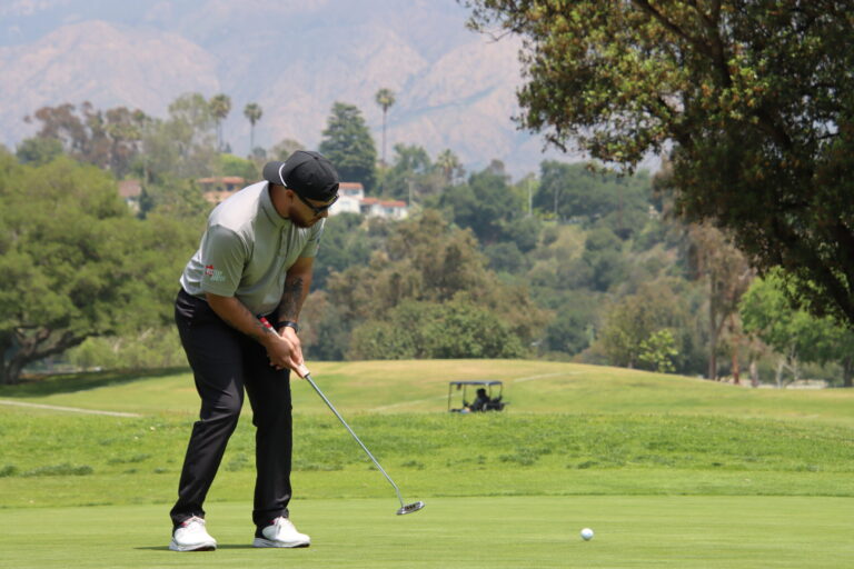 a man in black pants and a gray shirt wearing a backwards black ballcap putts a ball towards a hole