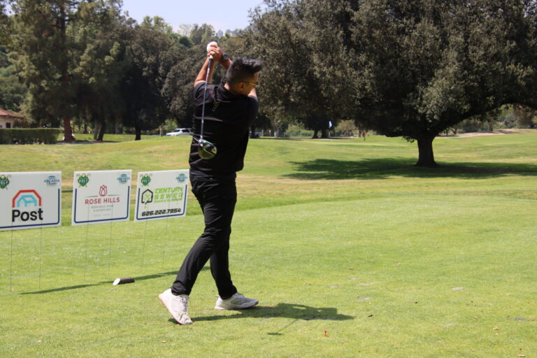 a man in a black shirt and pants with white sneakers tees off in front of a row of tee signs