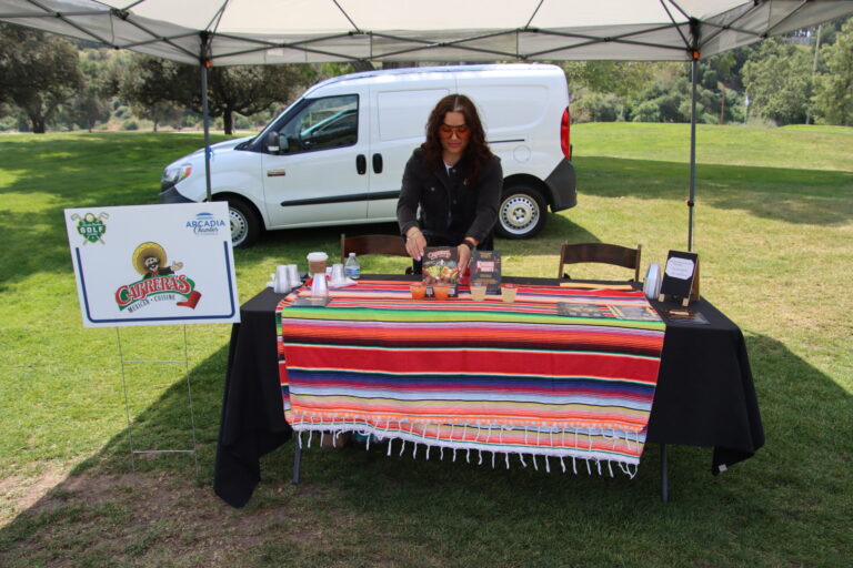 a woman standing behind a table set with a colorful table cloth, with a white van behind her, arranges margaritas on the table for Cabrera's