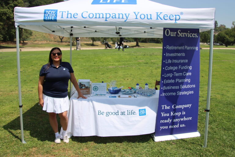 a woman in a blue shirt and white skirt poses in front of a tent for New York Life set with NYF items and a banner
