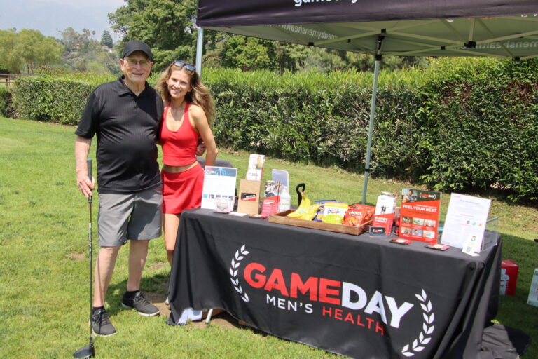 a woman in a red tank top and skirt posing with an older man in black and gray beside a table set with Gameday Men's Health items
