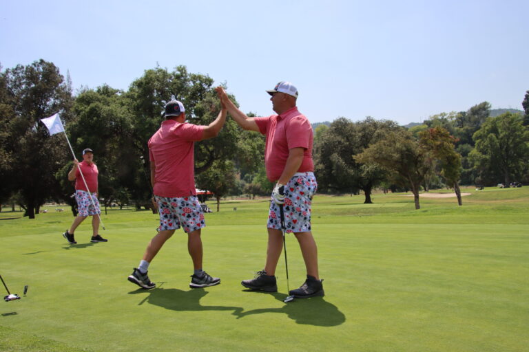 two men pink shirts and colorful shorts high fiving on a golf green while a similarly dressed man behind them holds a flag