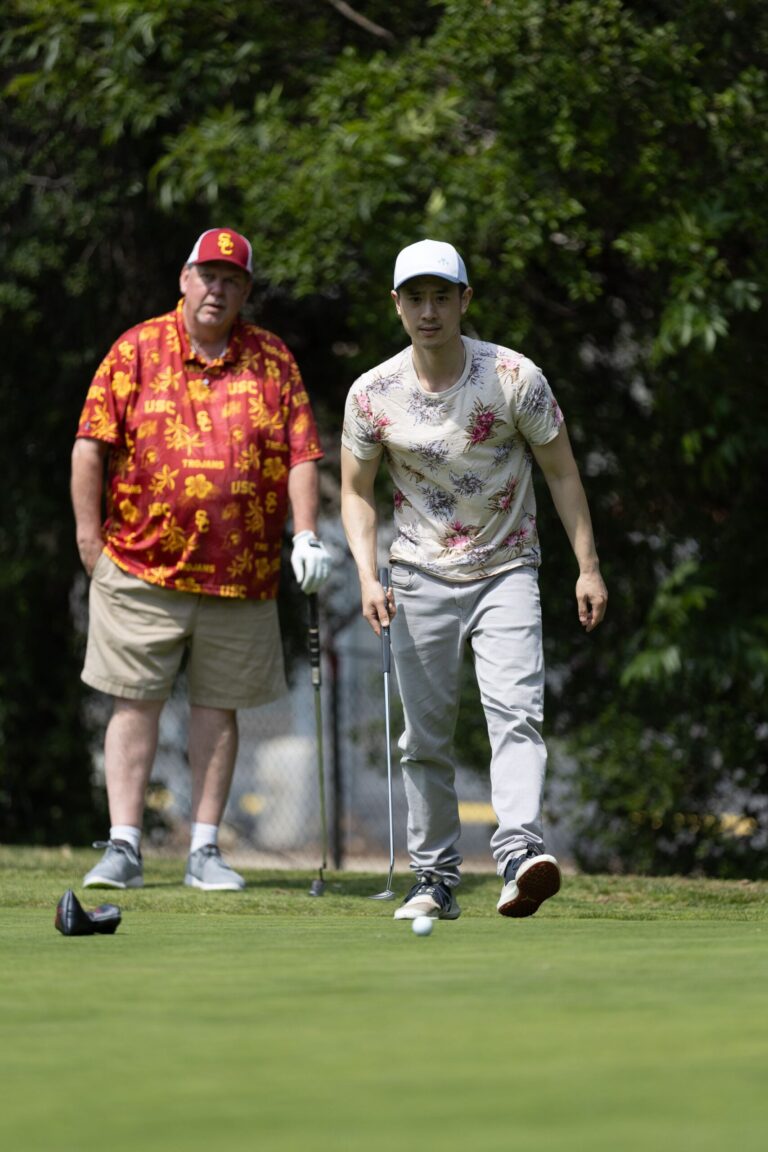 a man in a patterned red and gold shirt with another man in a patterned shirt walk towards a golf ball on a golf green