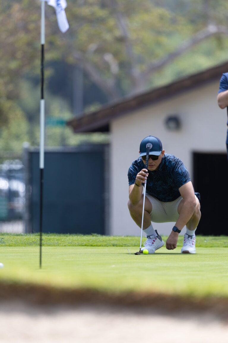 a man in a blue shirt and khaki shorts crouching down to look at a golf green with the flag in the foreground
