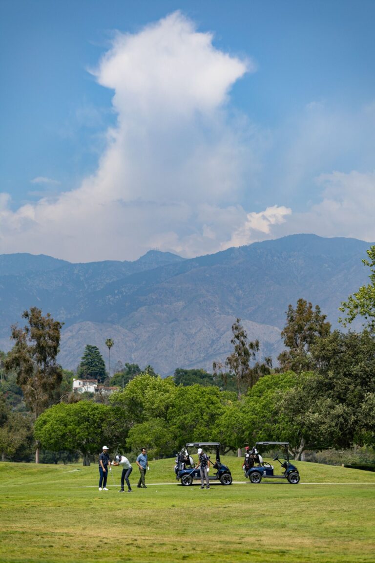 a group of golfers alongside two golf carts with the mountains, blue skies and large white clouds behind them