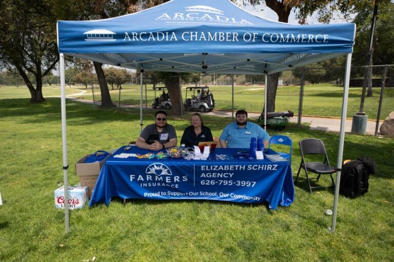 three people sit behind a booth with a blue tent and a blue Farmers Insurance table cloth