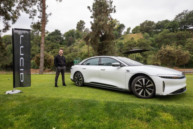 a man in black stands near a white Lucid sedan in a lush green setting
