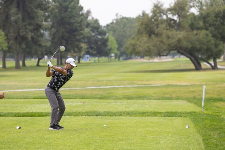a man in a black patterned shirt and gray pants swings at a golf ball on a golf course