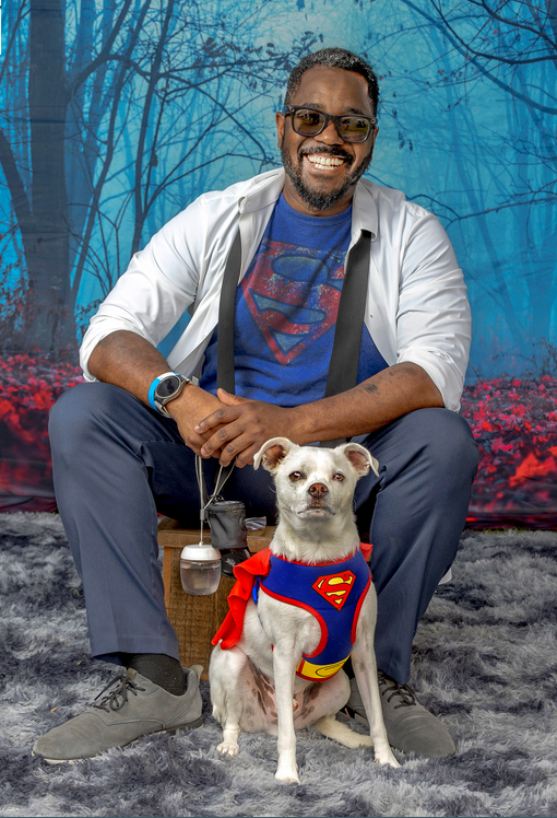a man in a superman shirt seated with a white dog in a superman costume