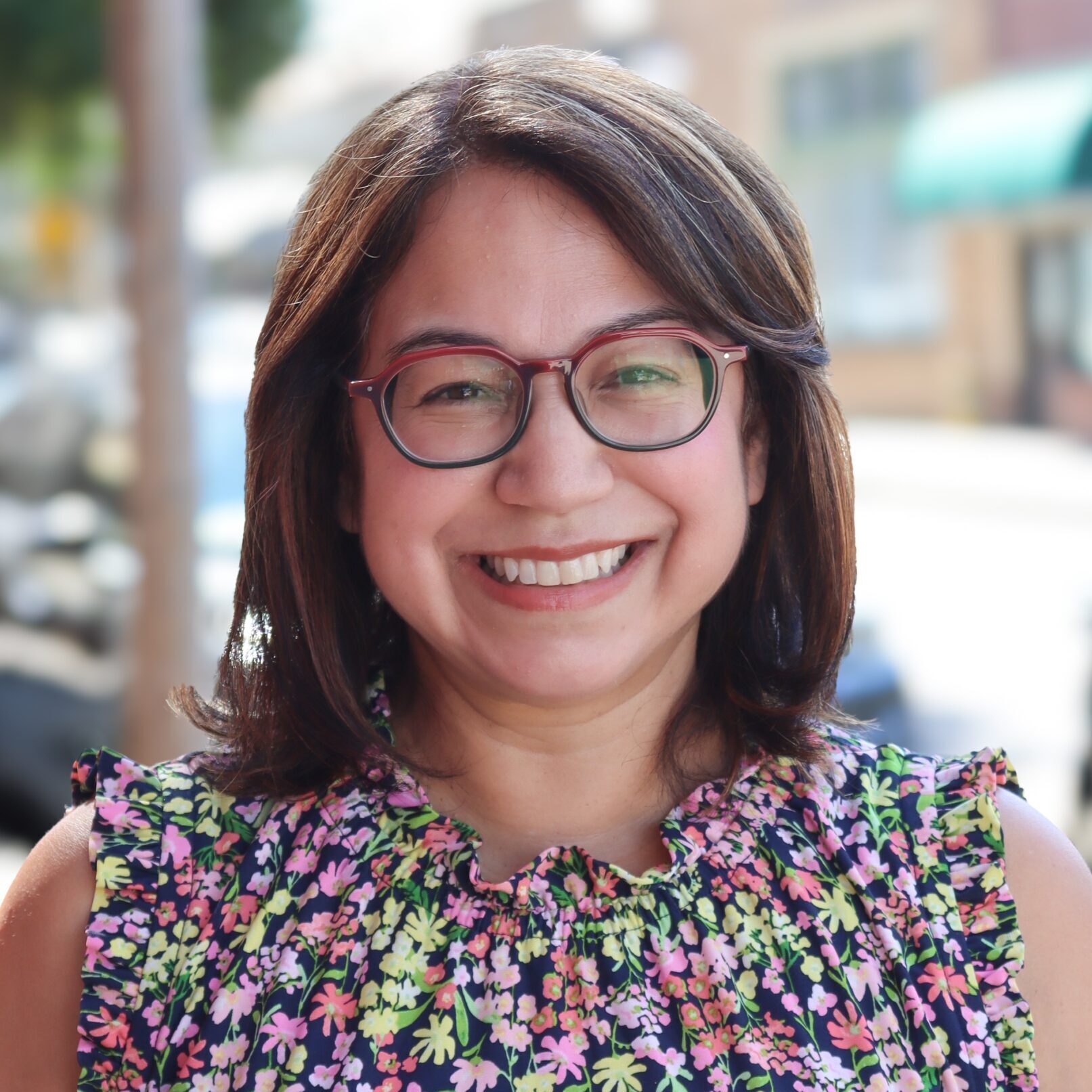 headshot for Monica Valenzuela, who is dressed in a floral print dress and wearing glasses