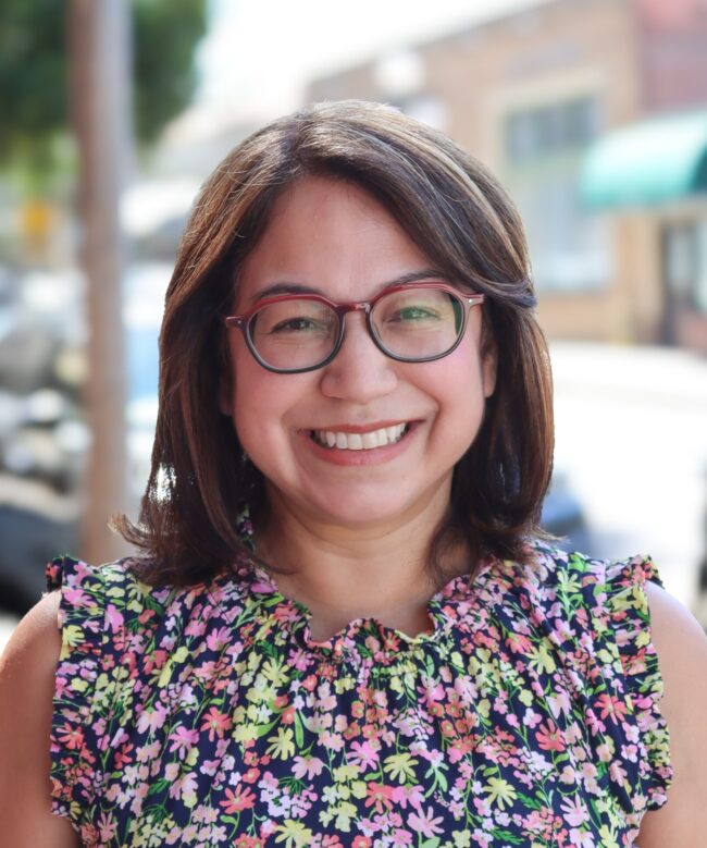 headshot for Monica Valenzuela, who is dressed in a floral print dress and wearing glasses