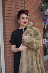 a woman in a black dress holds a fur coat while leaning on a brick wall