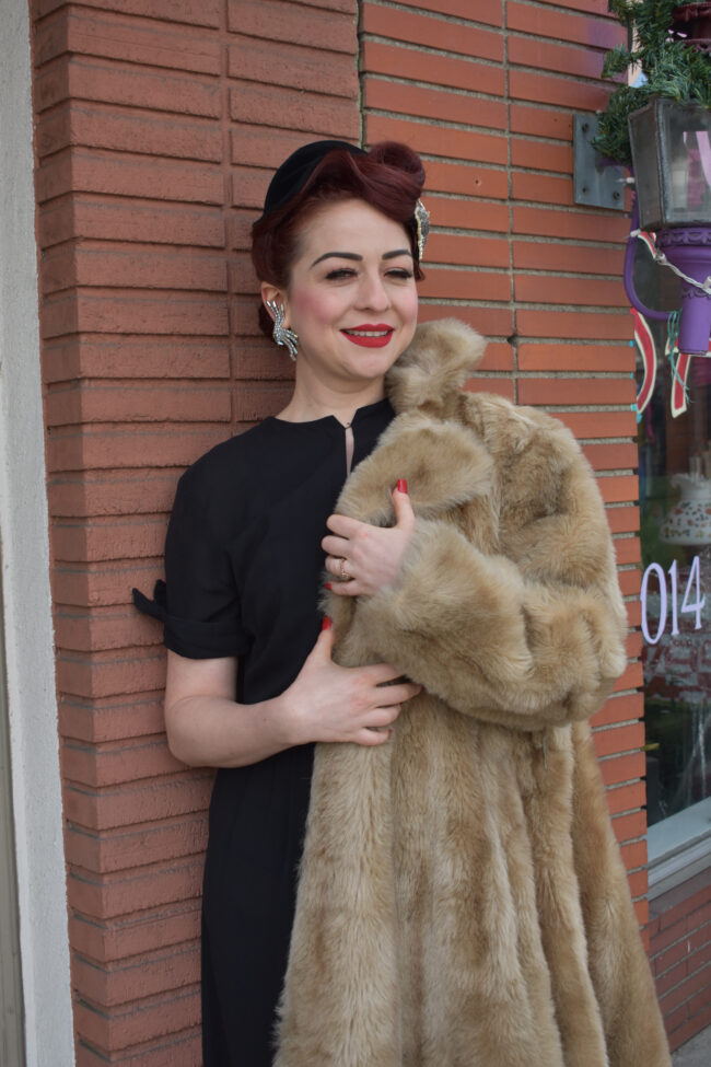 a woman in a black dress holds a fur coat while leaning on a brick wall