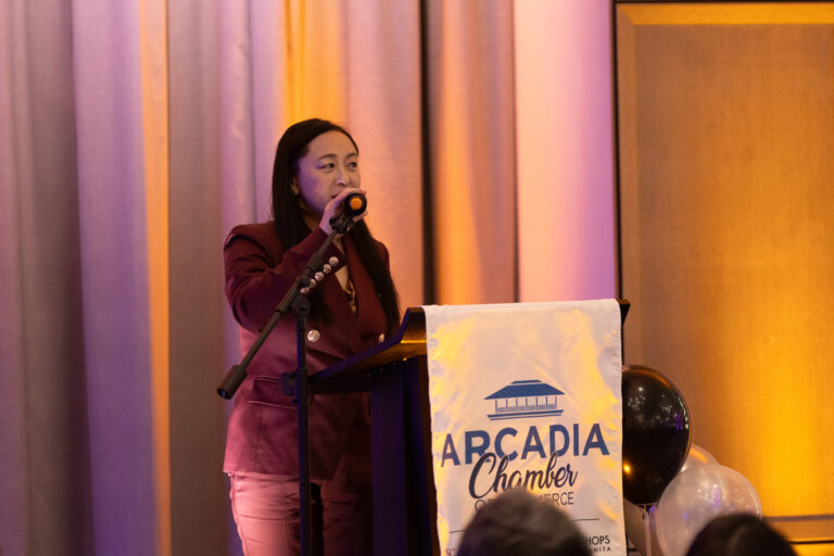 a woman in a purple suit standing behind a podium set with the Arcadia Chamber logo