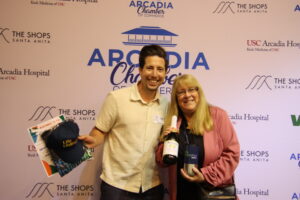 IMG_4170 a woman in a pink shirt with a taller man in a white shirt standing in front of a step and repeat with the Arcadia Chamber logo on it