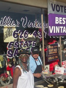 a woman with long gray hair in braids and wearing a hat standing in front of a man wearing a blue shirt and apron who is standing over a grill top