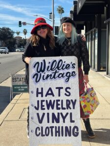two women wearing hats standing behind a sandwich board that says Willie's Vintage Hats, Jewelry and Vinyl
