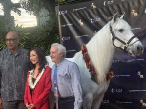 a woman in a red jacket with a man in a blue shirt and tie standing in front of a gray horse