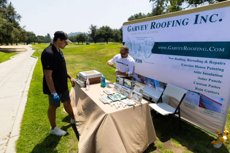 a man in black looks at a table of items while a man in a white shirt stands behind the table, with a Garvey Roofing step and repeat behind them
