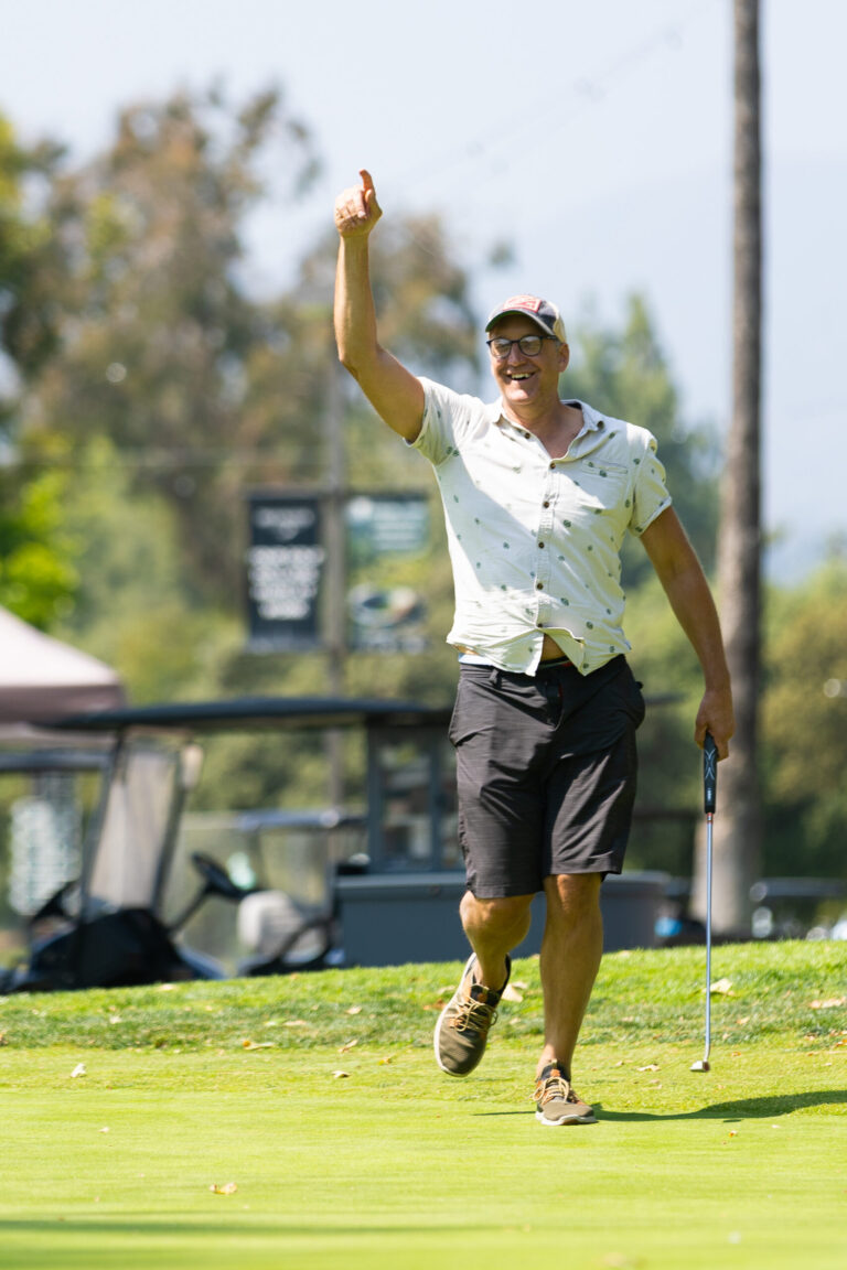 a man in a white golf shirt with black shorts holds a hand up in celebration while carrying a golf club