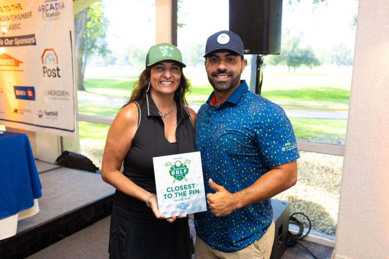 a woman in a black dress and green baseball hat stands beside a man in a blue shirt holding a plaque and giving a thumbs up