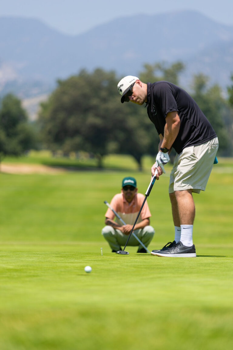 a man in a black shirt and white shorts putts a ball on a golf course while another man squats behind him watching