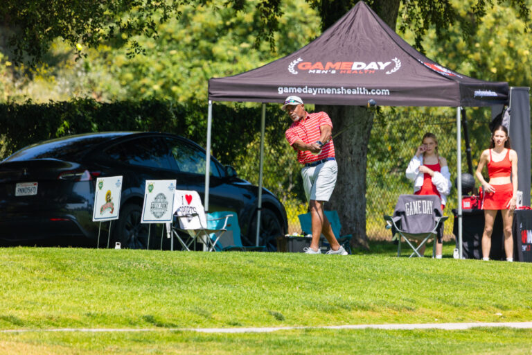 a man in a red shirt and white shorts hits a ball in front of a tent labeled with the Gameday Men's Health logo