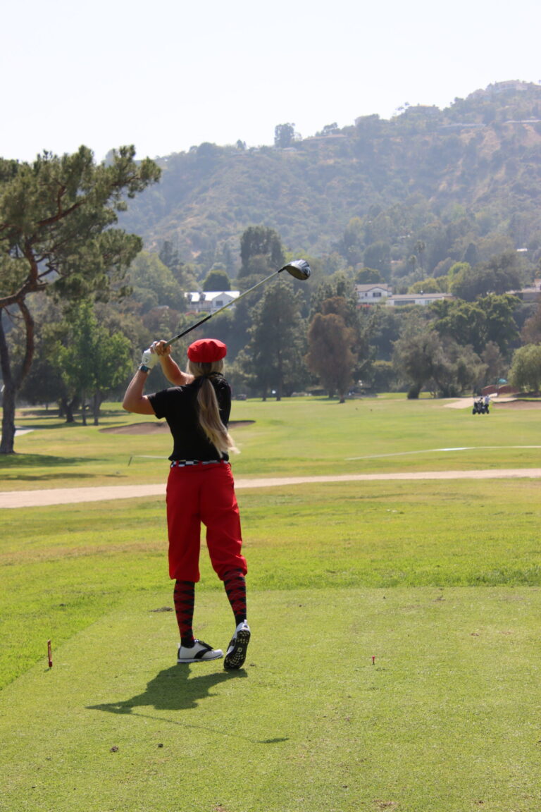 a female golfer in red pants and newsboy cap driving a ball on the fairway