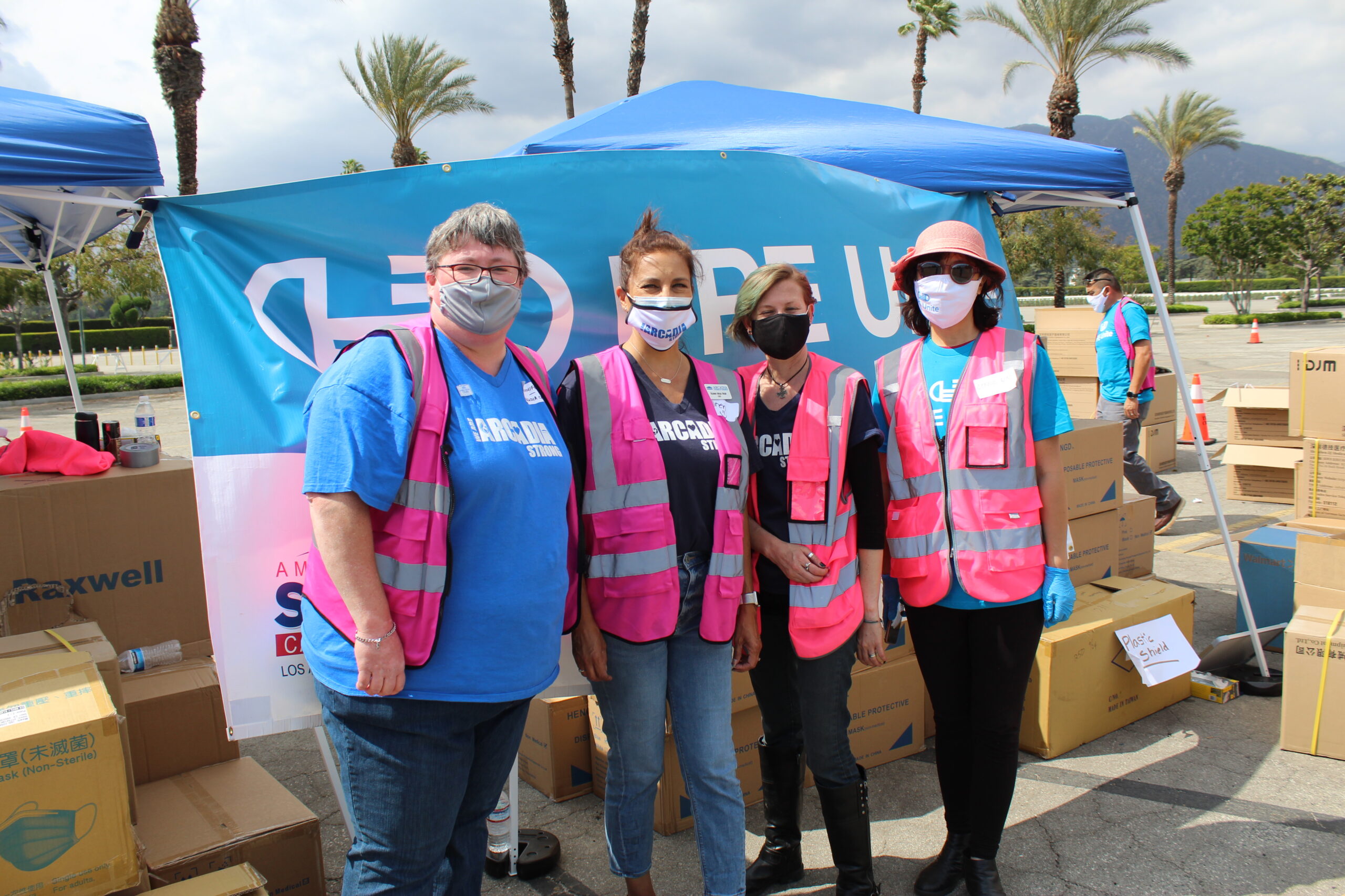 IMG_6712 a group of four women standing in front of a blue banner, all wearing pink reflective vests and masks